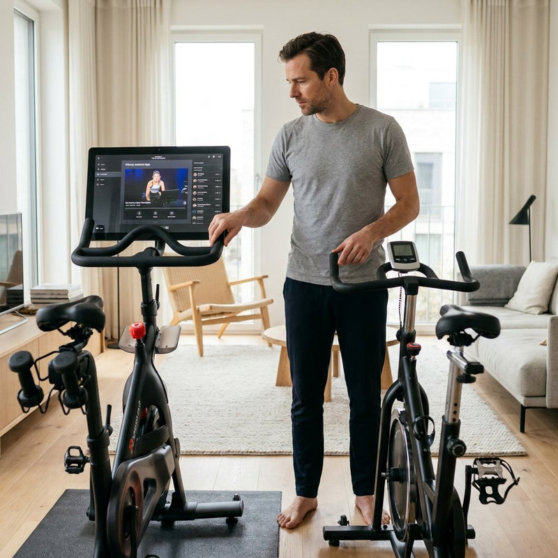 Moderne thuissporter test twee spinningfietsen in een lichte woonkamer: één met groot scherm en één met eenvoudig display, in een rustige thuissetting.